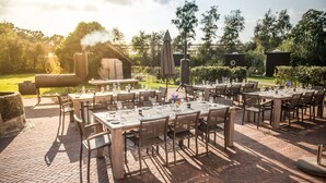 An outdoor dining area with brick flooring, wooden tables, and chairs. - Erfgoed Bossem (Lattrop)