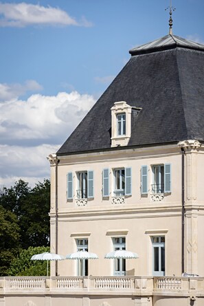 A building with a steeply pitched roof, multiple windows with shutters, and a balcony with umbrellas. - Château de Cîteaux - Fontenille Collection (Meursault)