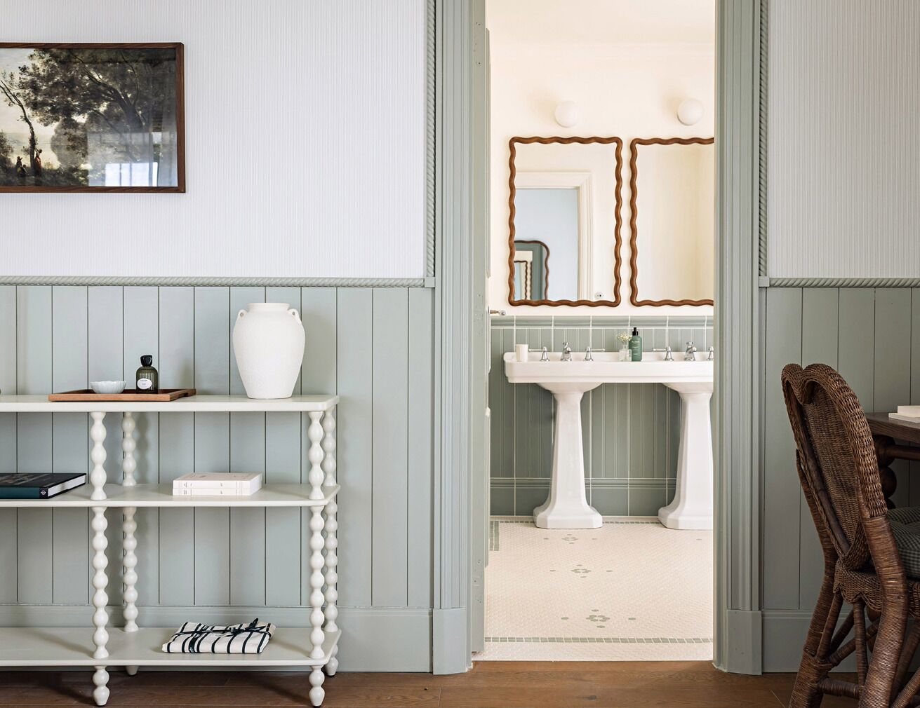 A room with a white pedestal sink, a mirror with a wavy frame, and a wicker chair.