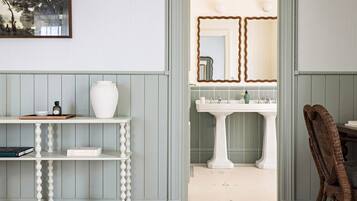 Une pièce avec un lavabo sur pied blanc, un miroir à cadre ondulé et une chaise en osier.