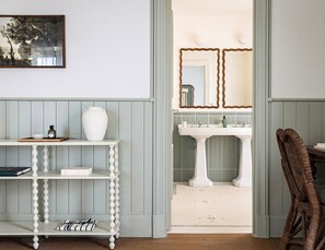 A room with a white pedestal sink, a mirror with a wavy frame, and a wicker chair. - Château de Cîteaux - Fontenille Collection (Meursault)