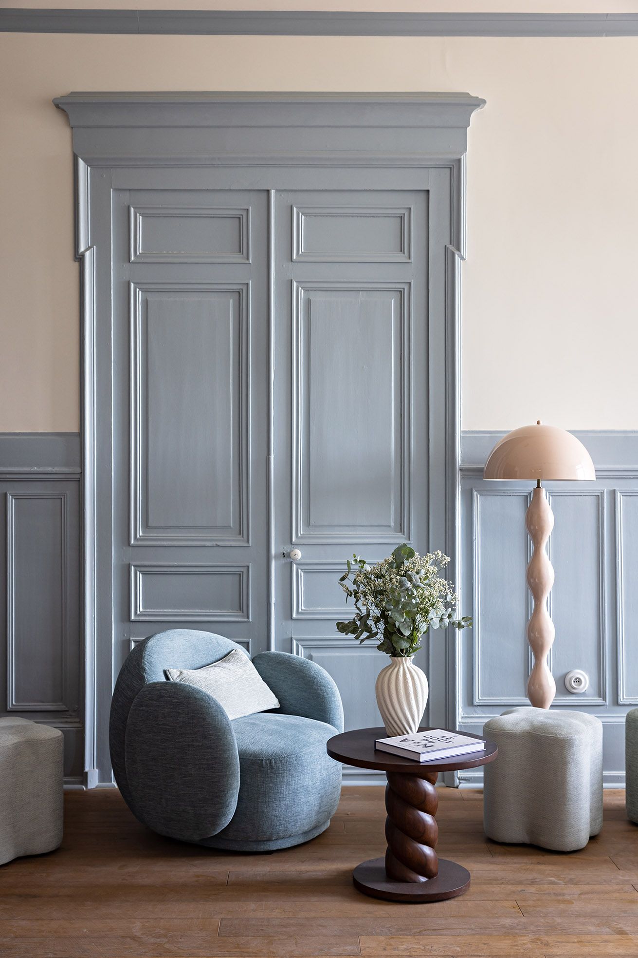 A room with a grey upholstered armchair, a small round table with a vase of flowers, and a floor lamp.