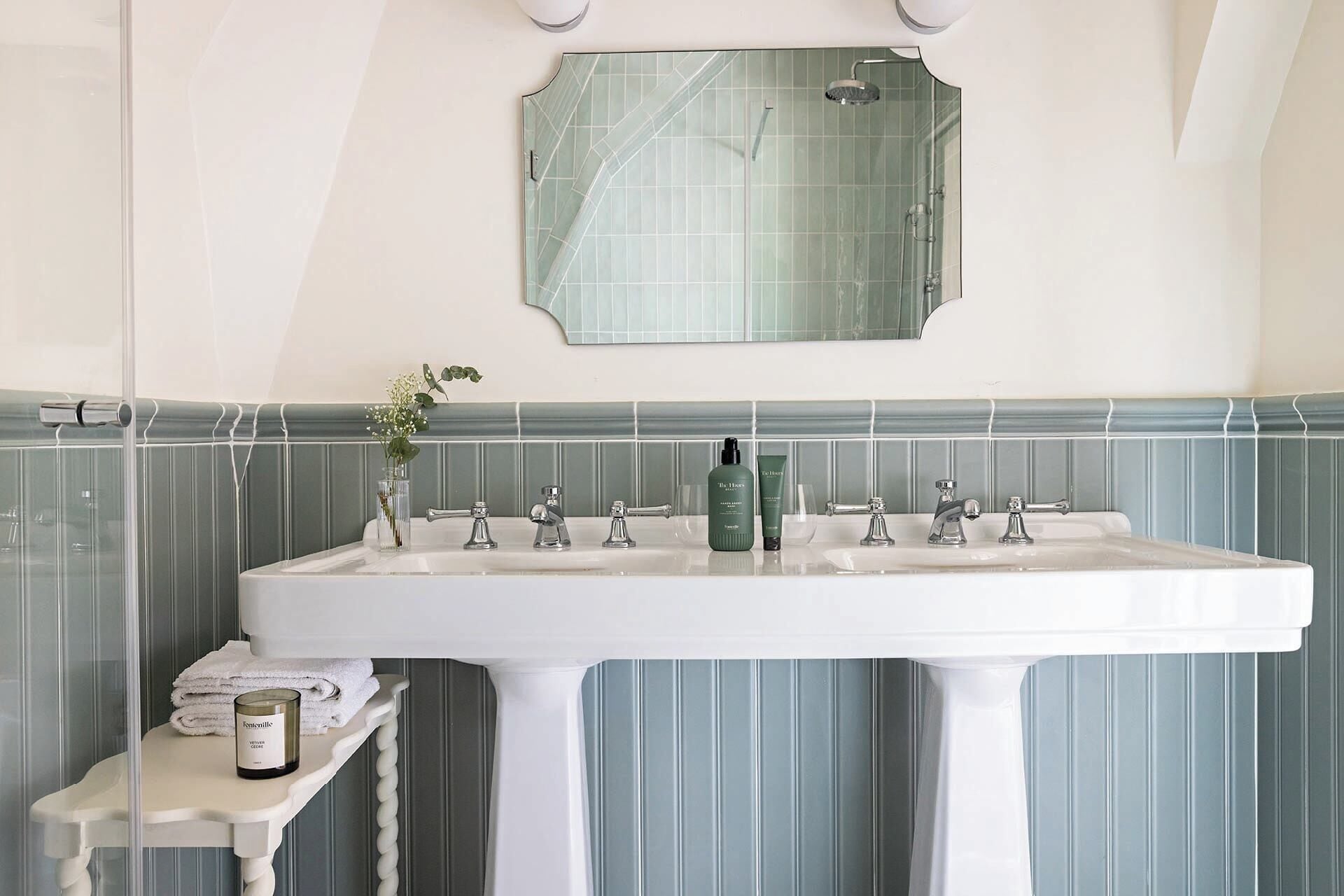 A bathroom with a large white sink, two faucets, a mirror, and a small table with a candle and a glass of flowers.