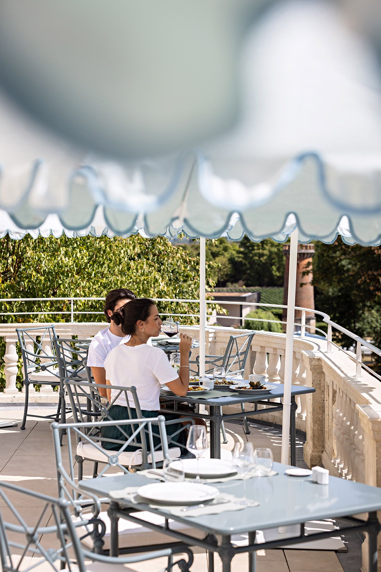 Een terras met tafels en stoelen, een witte parasol en uitzicht op groen.