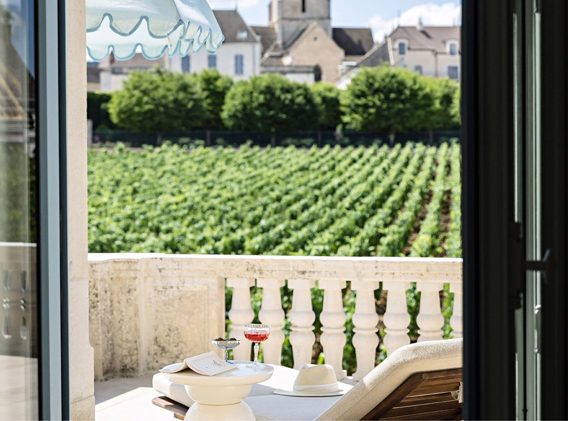 A balcony with a view of a vineyard, a table with a hat, and a bottle.