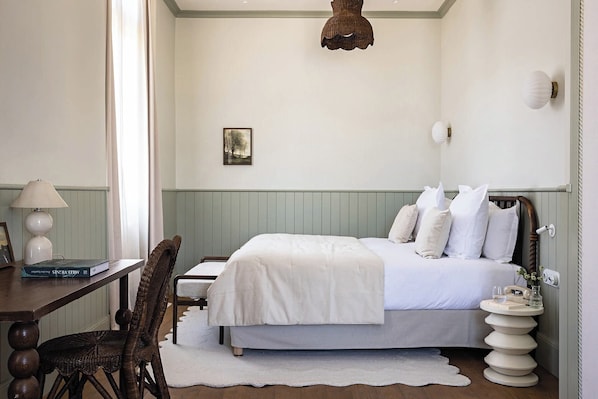 Een slaapkamer met een bed, nachtkastje, lamp, stoel en een kleintje tafeltje met een boek.