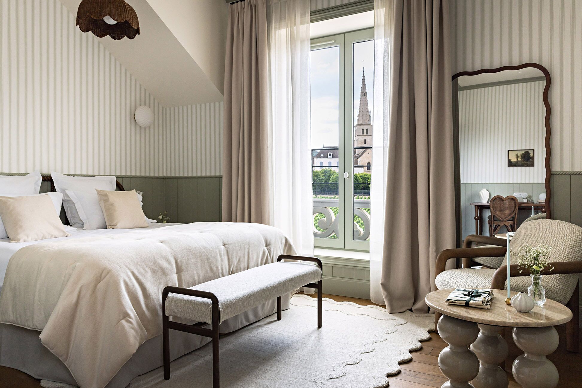 A hotel room with a bed, bench, round table, and a view of a building through the window.