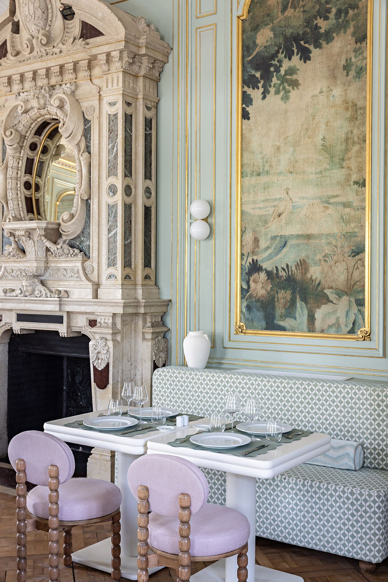 Un comedor con una mesa dispuesta para cuatro personas, una chimenea, decoraciones ornamentales en la pared y un cuadro de paisaje grande.