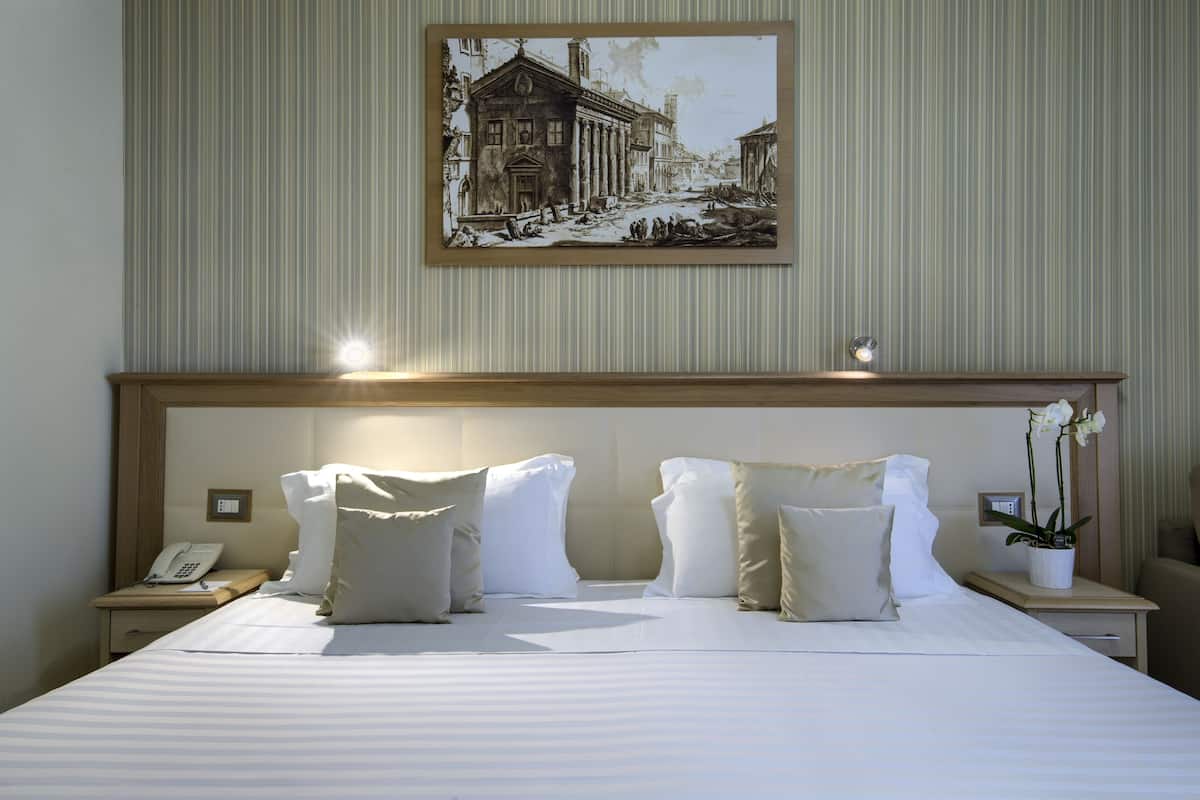 A neatly made bed with white linens and multiple pillows, a bedside telephone, and a small table with a potted orchid.