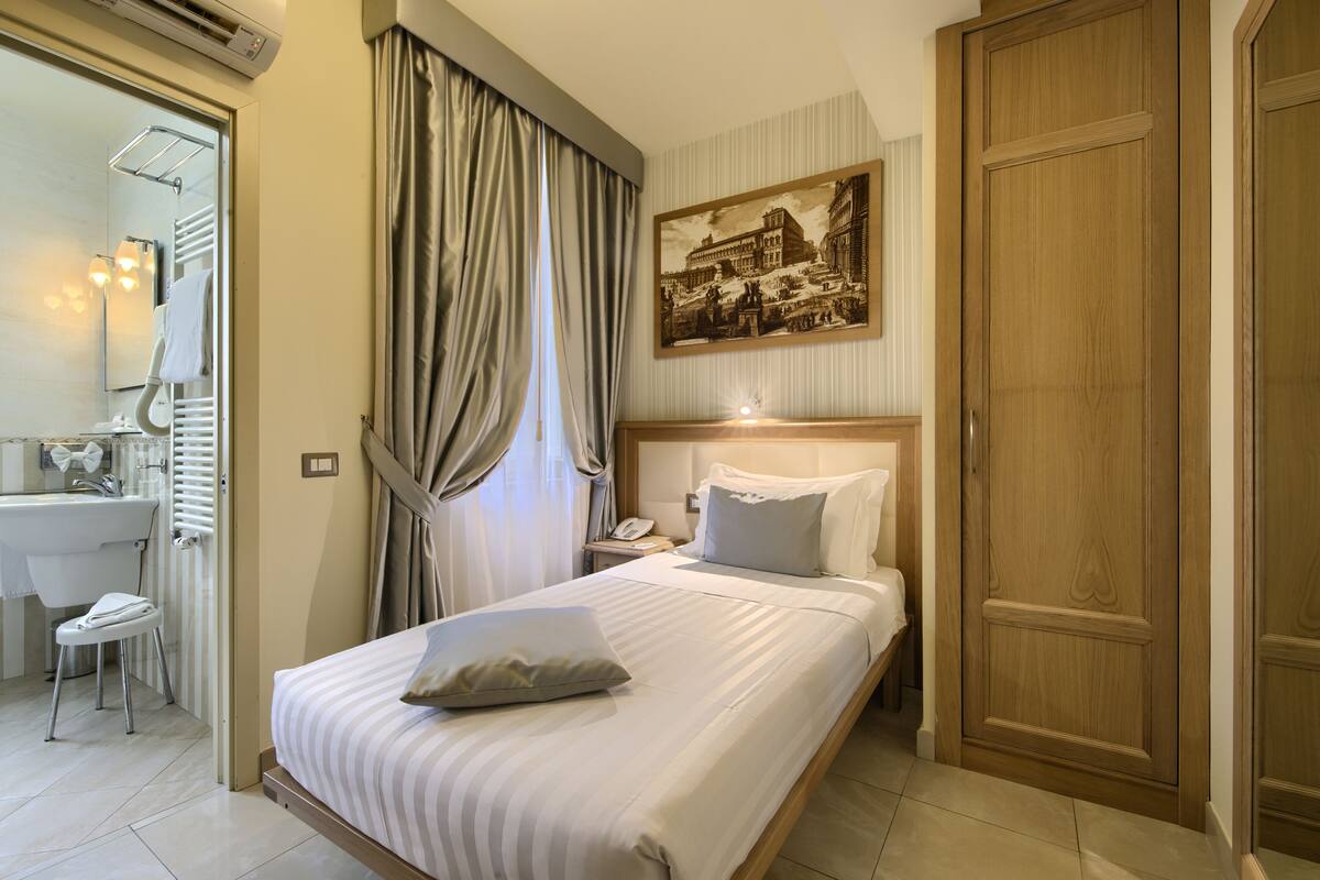 A hotel room with a bed, a nightstand, a bedside table, a mirror, a window with curtains, a bathroom with a sink and toilet, and a door.