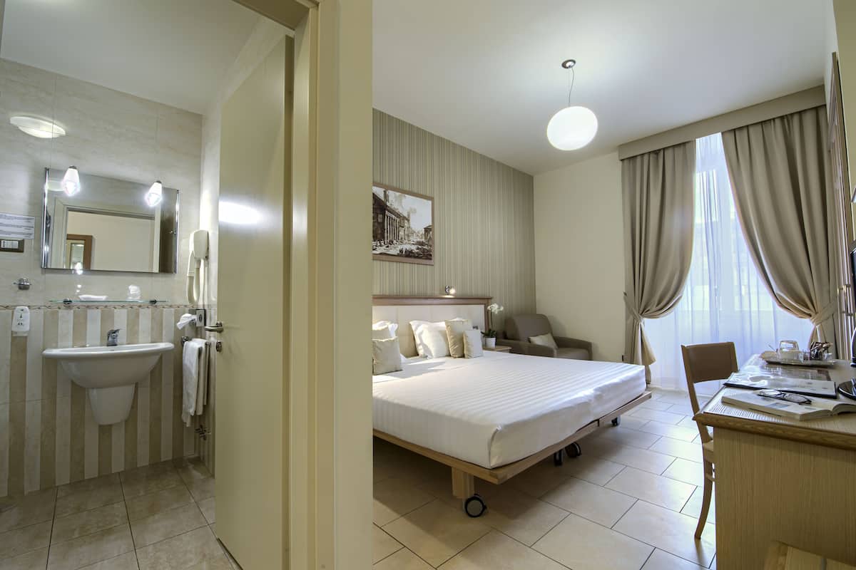 A hotel room with a bed, bedside table, chair, desk, and a bathroom with a sink and mirror.