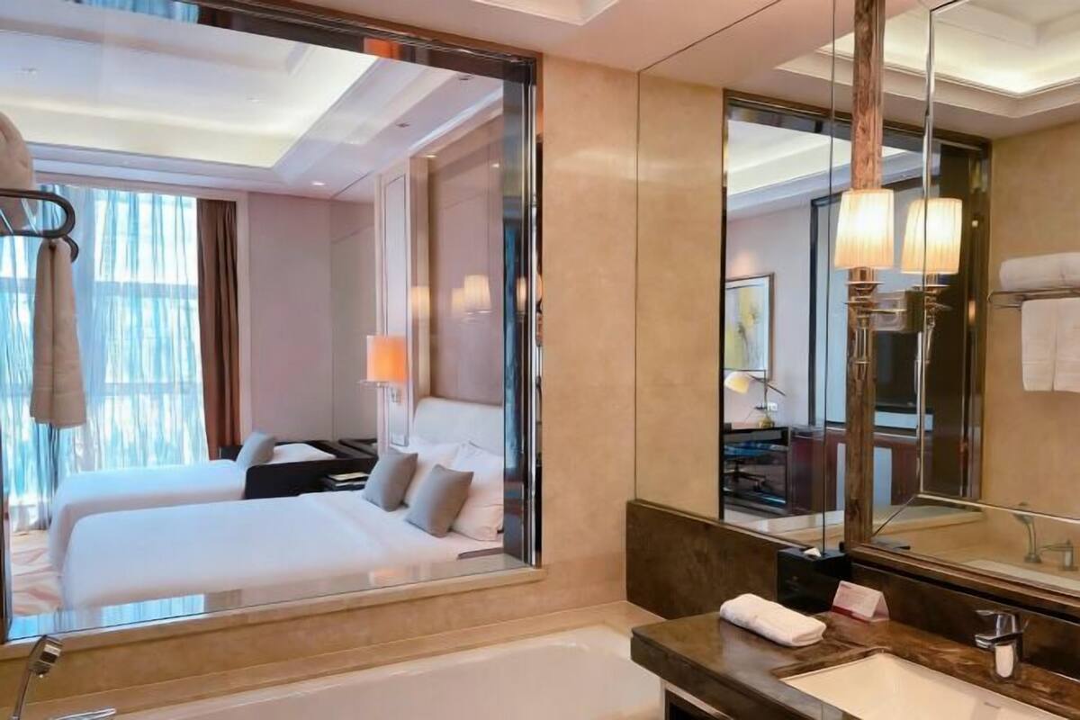 Quarto de hotel com um espelho grande, uma banheira, uma pia e duas camas.
