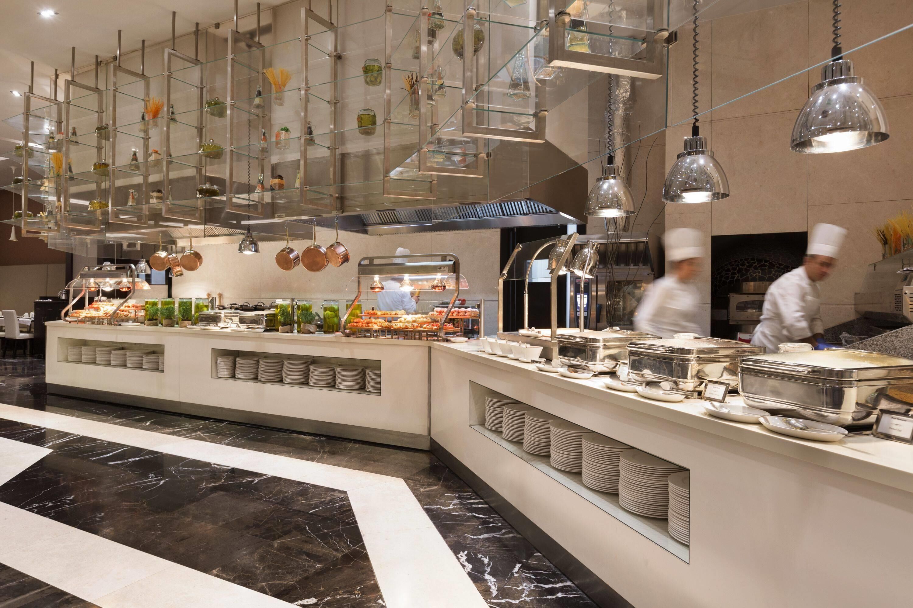 Uma área de buffet com várias estações de serviço, prateleiras de vidro com objetos decorativos e chefs preparando alimentos.