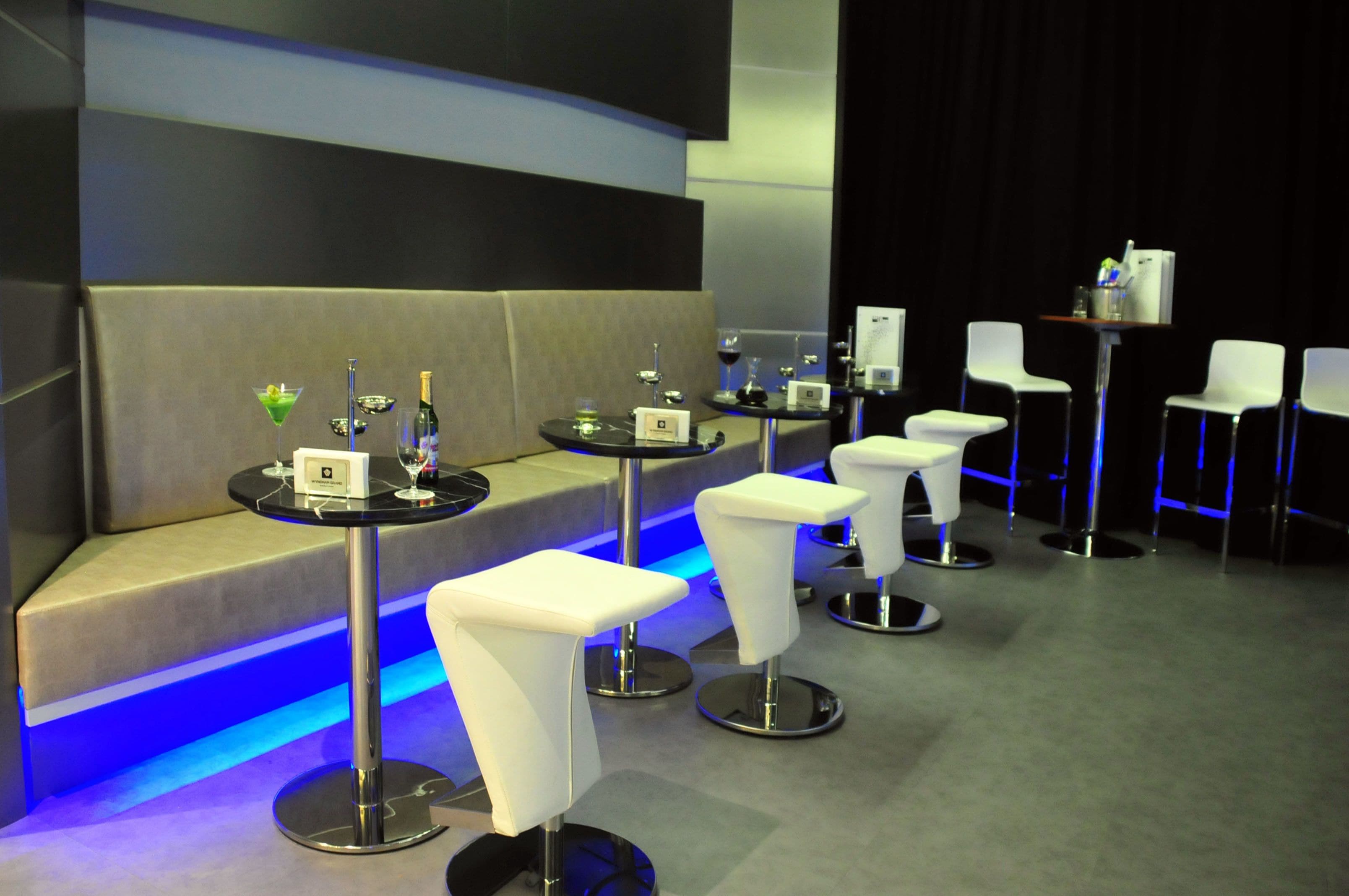 A modern lounge area with a sofa and bar stools, a small bar table with a bottle and glasses, and a stage with chairs.