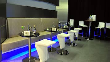 A modern lounge area with a sofa and bar stools, a small bar table with a bottle and glasses, and a stage with chairs.