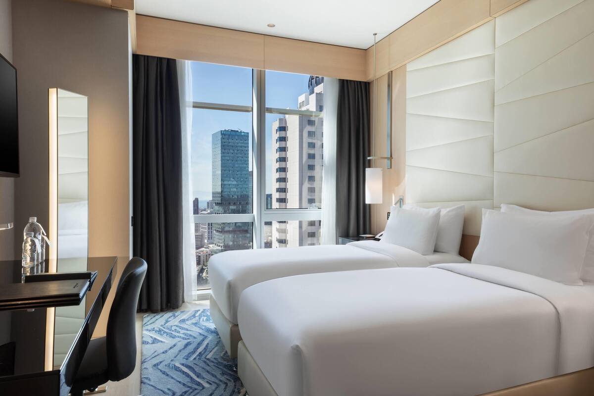 Ein Hotelzimmer mit zwei Betten, einem Schreibtisch, einem Stuhl, einem Fernseher und einem großen Fenster mit Blick auf die Stadt.