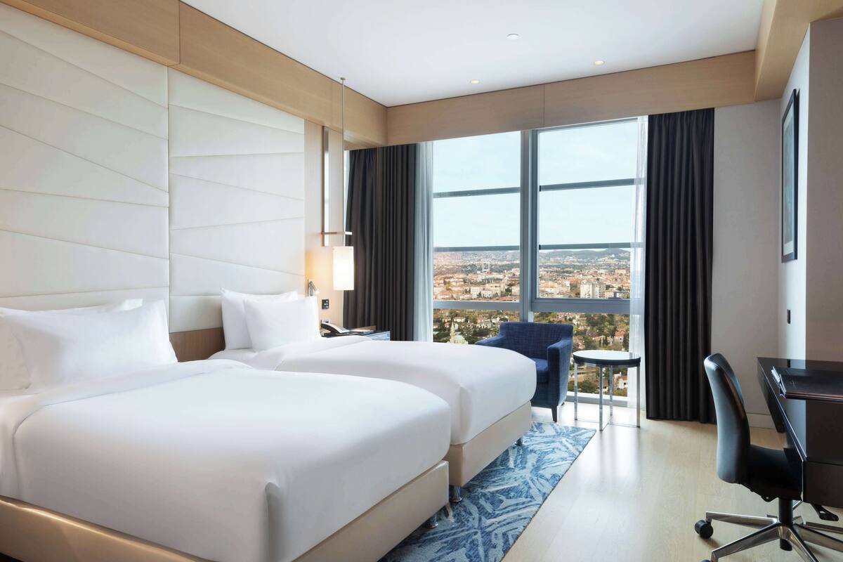 Ein Hotelzimmer mit einem großen Bett, einem Schreibtisch und Blick auf eine Stadtkulisse durch große Fenster.