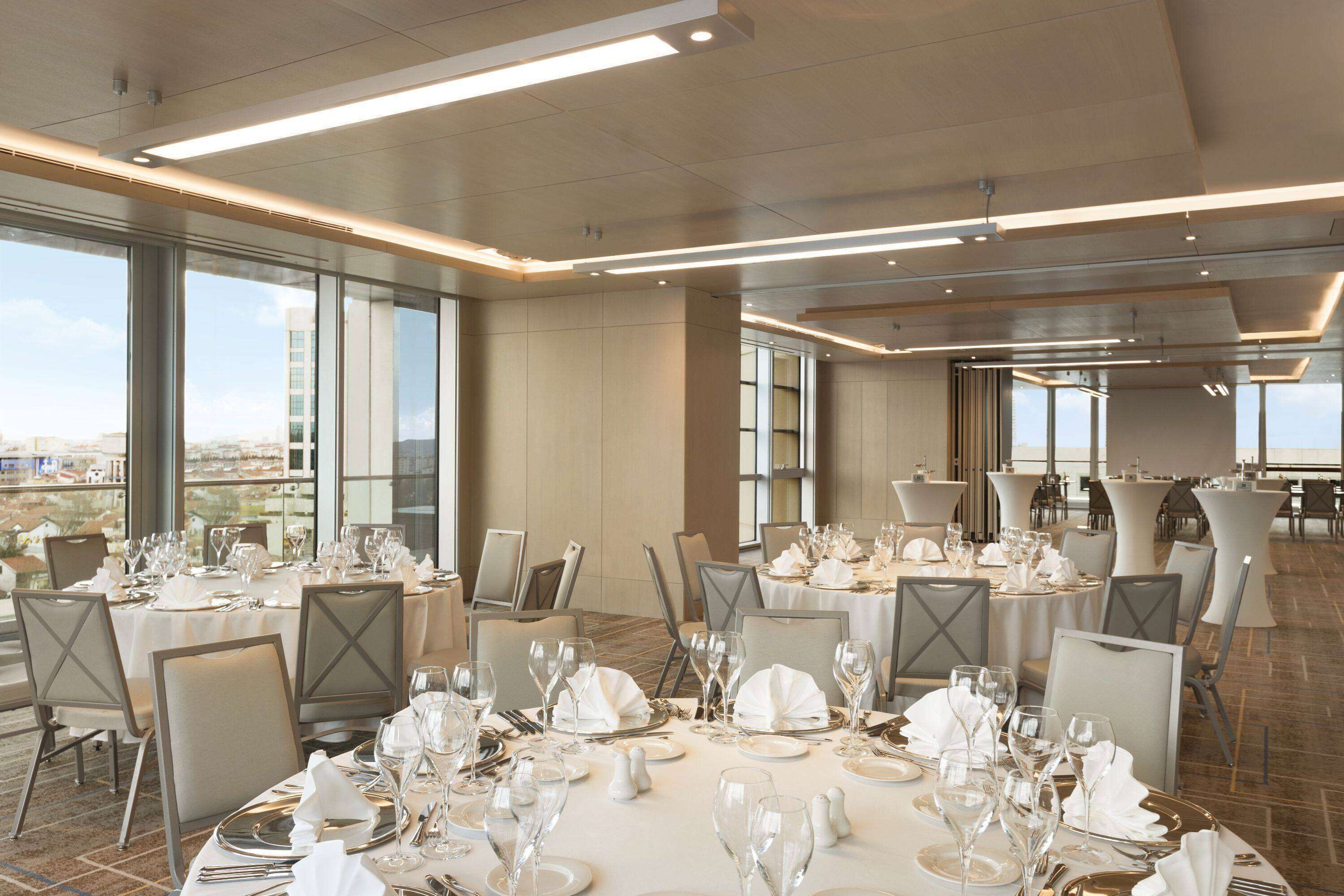 A spacious dining area with multiple tables set for a formal meal, large windows offering a city view, and a modern ceiling design.