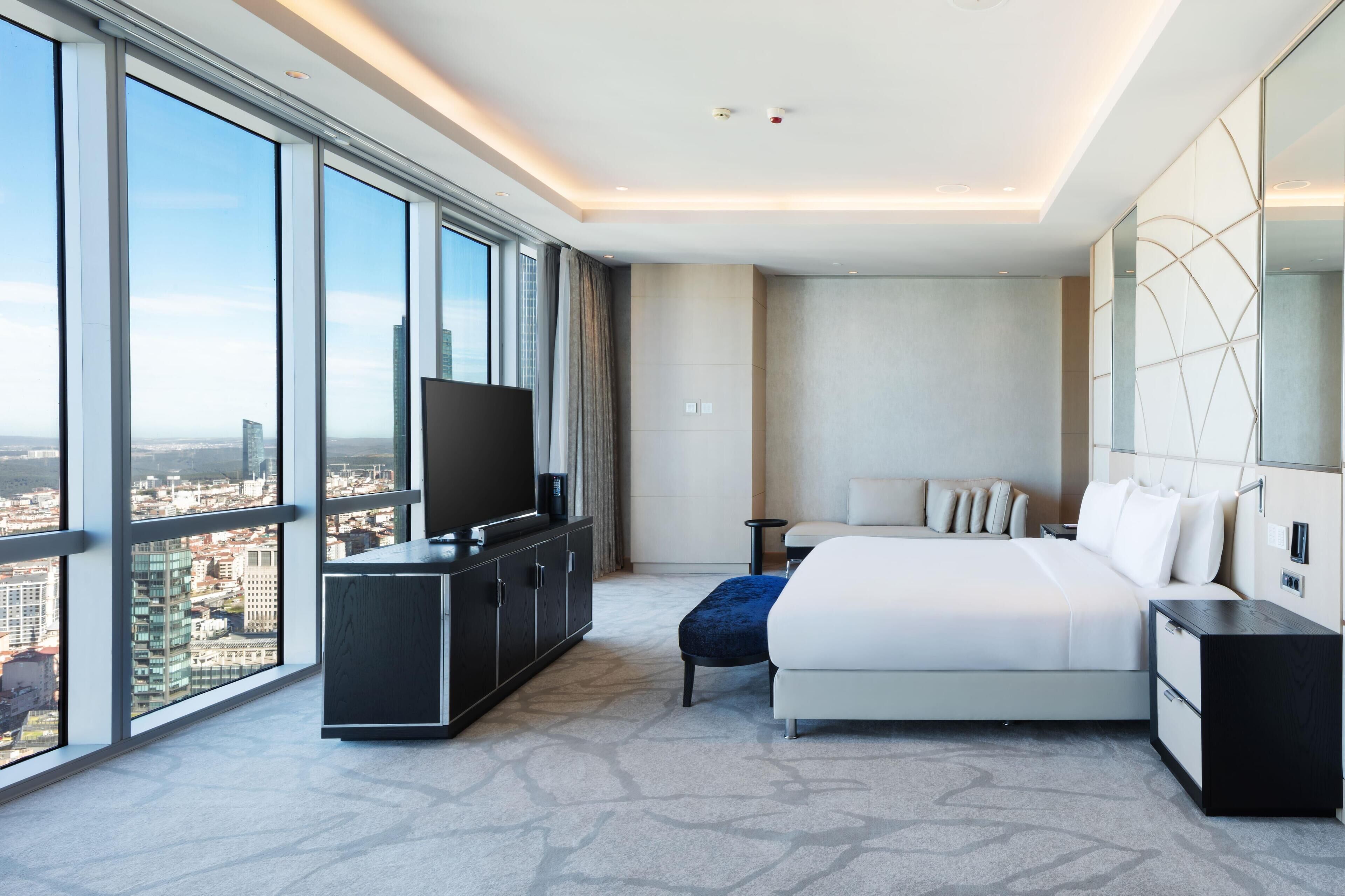 Quarto de hotel moderno com uma cama grande, uma TV em um móvel e vista para a cidade.
