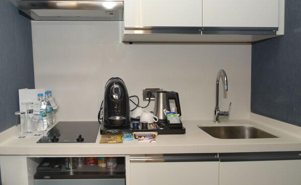 Una kitchenette con cafetera, hervidor de agua y pileta. Hay botellas de agua, un cápsula de café y una caja de snacks en la encimera.