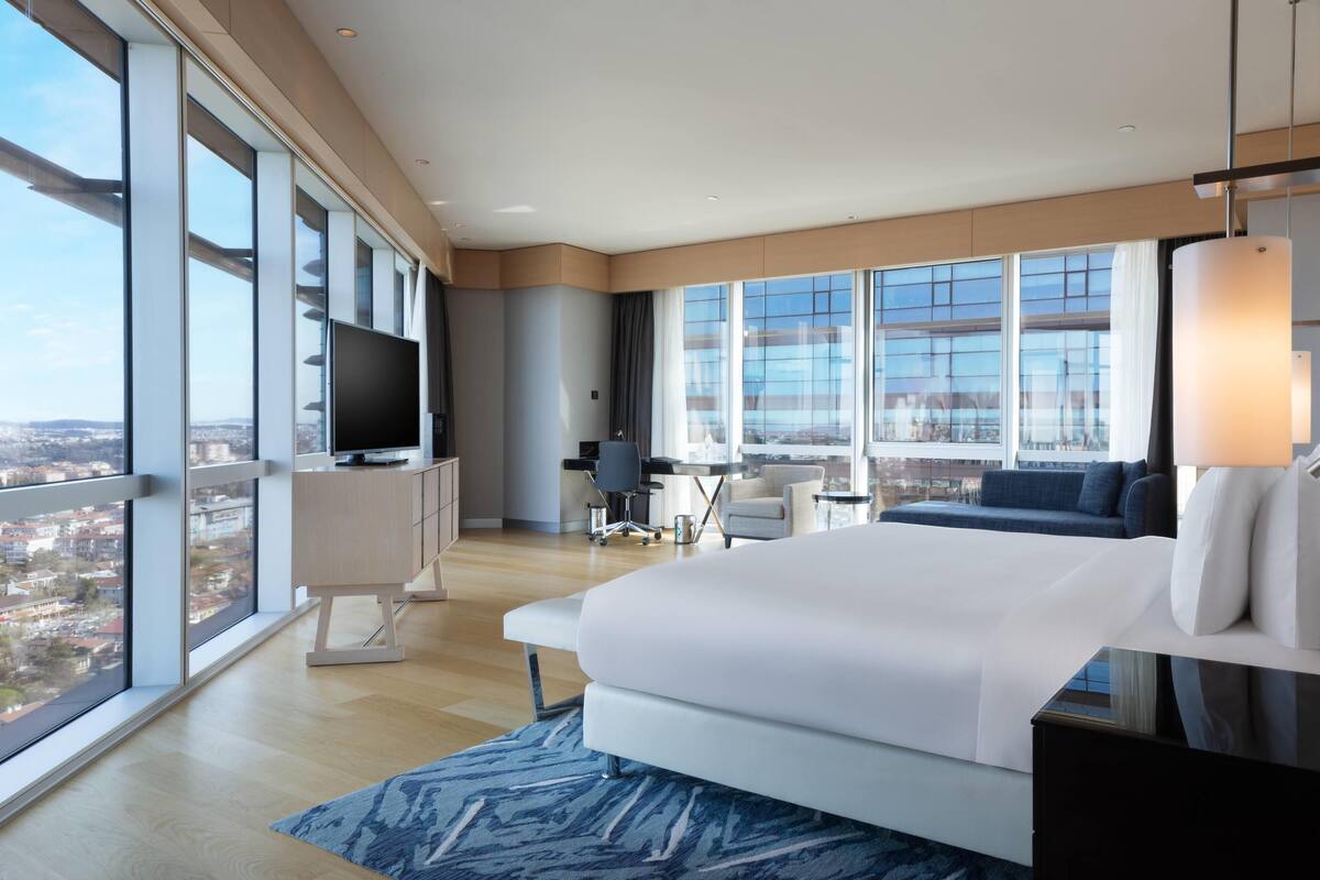 Ein modernes Hotelzimmer mit einem großen Bett, einem Schreibtisch, einem Sessel, einem Fernseher und Blick auf die Stadt.