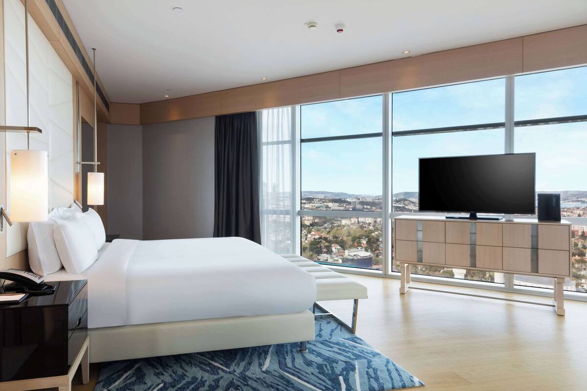 Ein modernes Hotelzimmer mit einem großen Bett, einem Flachbildfernseher, einem Schreibtisch mit Telefon und Blick auf die Stadt durch große Fenster.