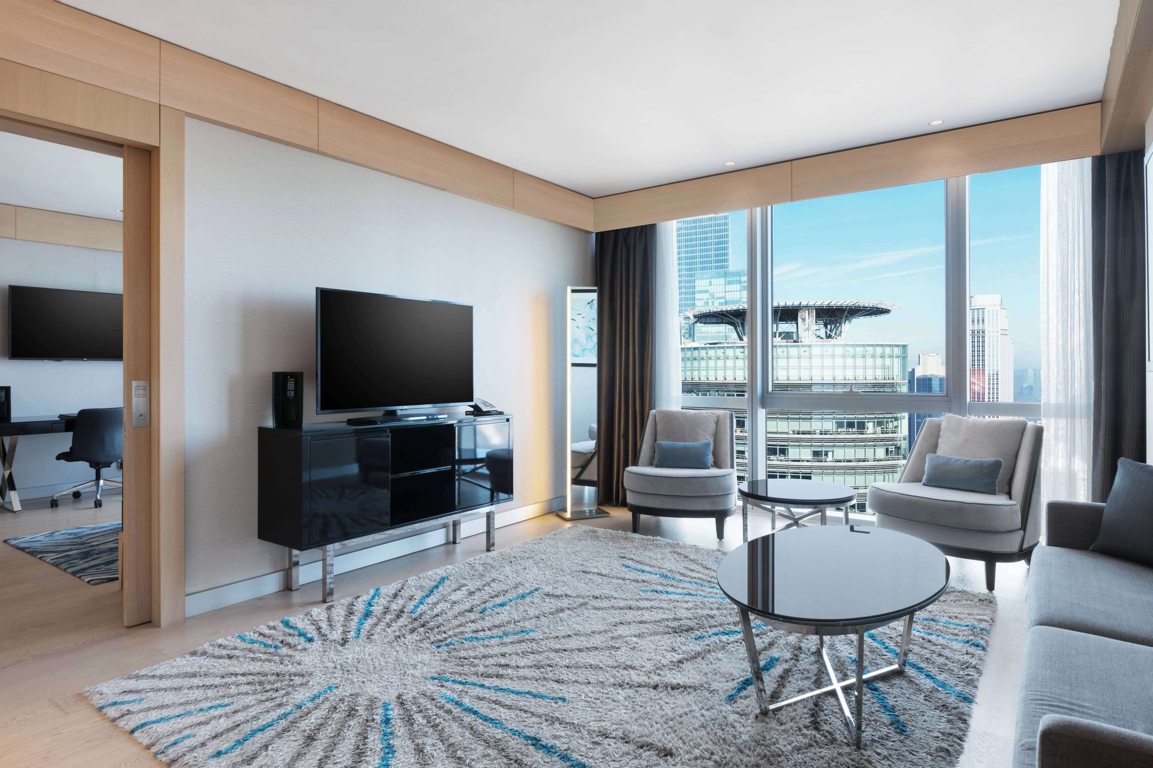 A modern hotel room with a large TV, comfortable seating, a view of the city, and a stylish rug.