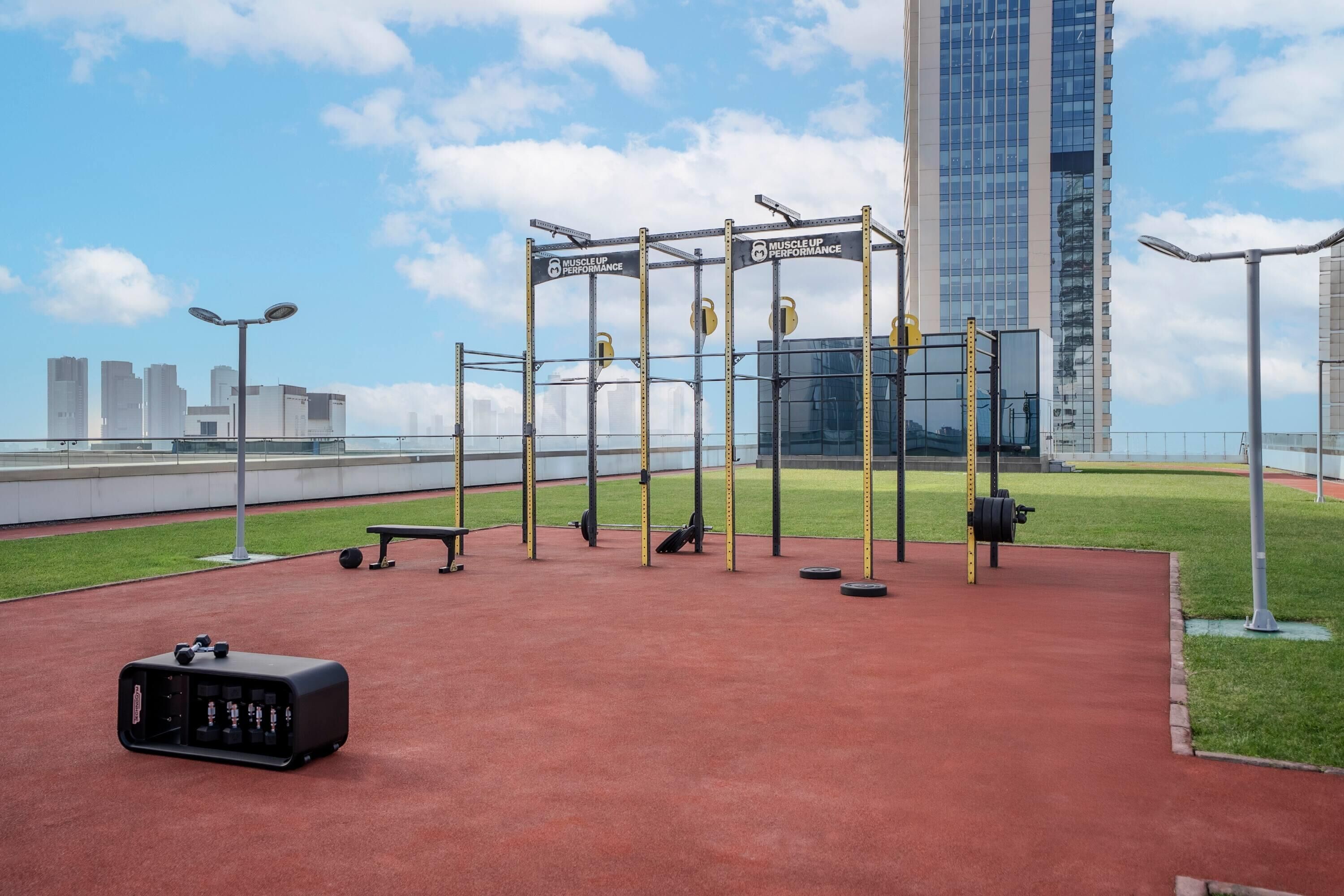An outdoor gym with weightlifting equipment, a bench, and a suitcase on a red surface.