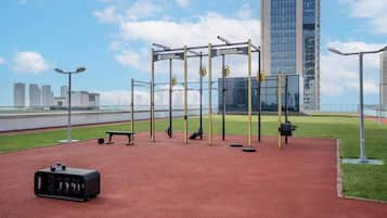 An outdoor gym with weightlifting equipment, a bench, and a suitcase on a red surface.