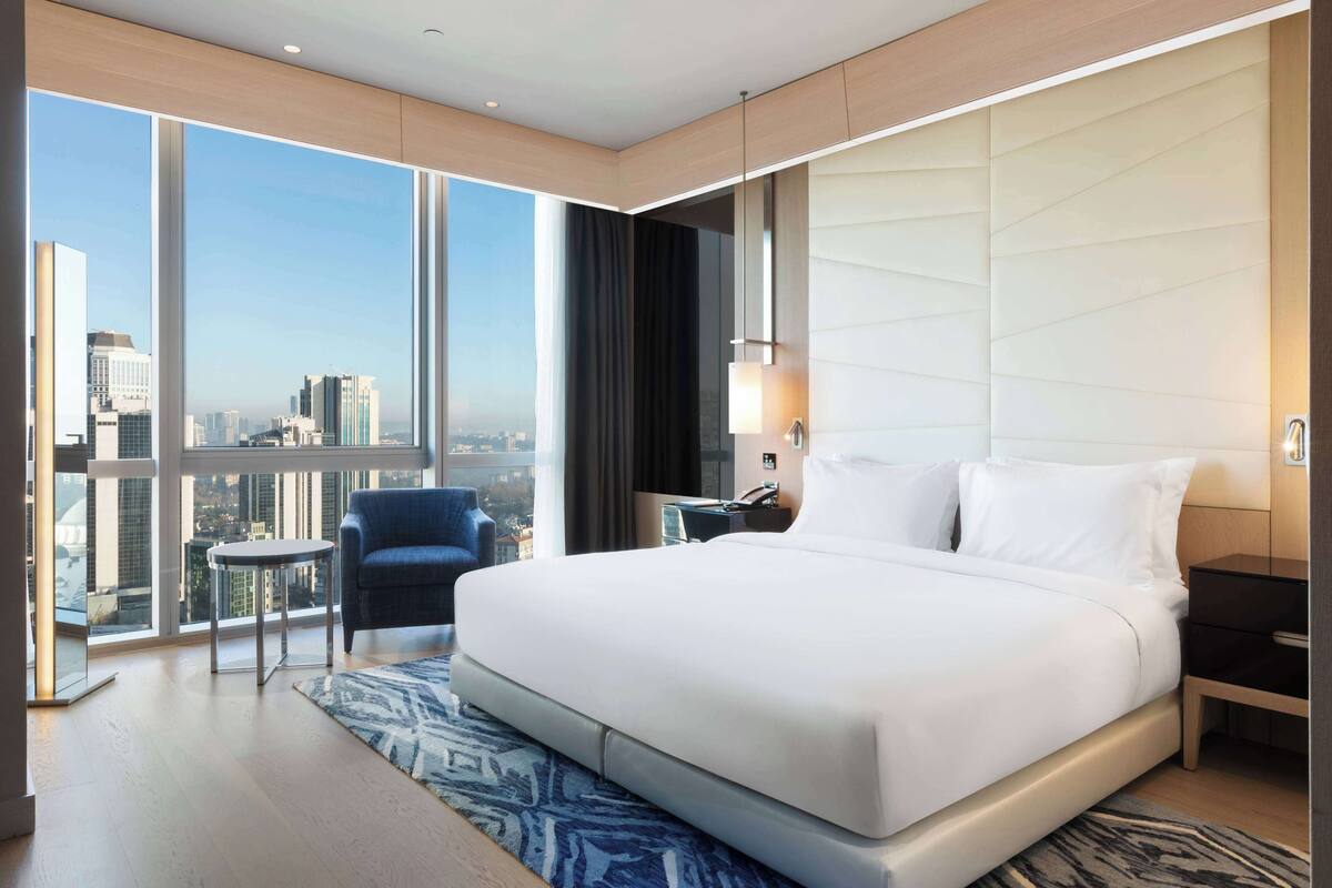 Ein modernes Hotelzimmer mit einem großen Bett, einem kleinen runden Tisch, einem blauen Sessel und einem Blick auf die Stadt durch große Fenster.