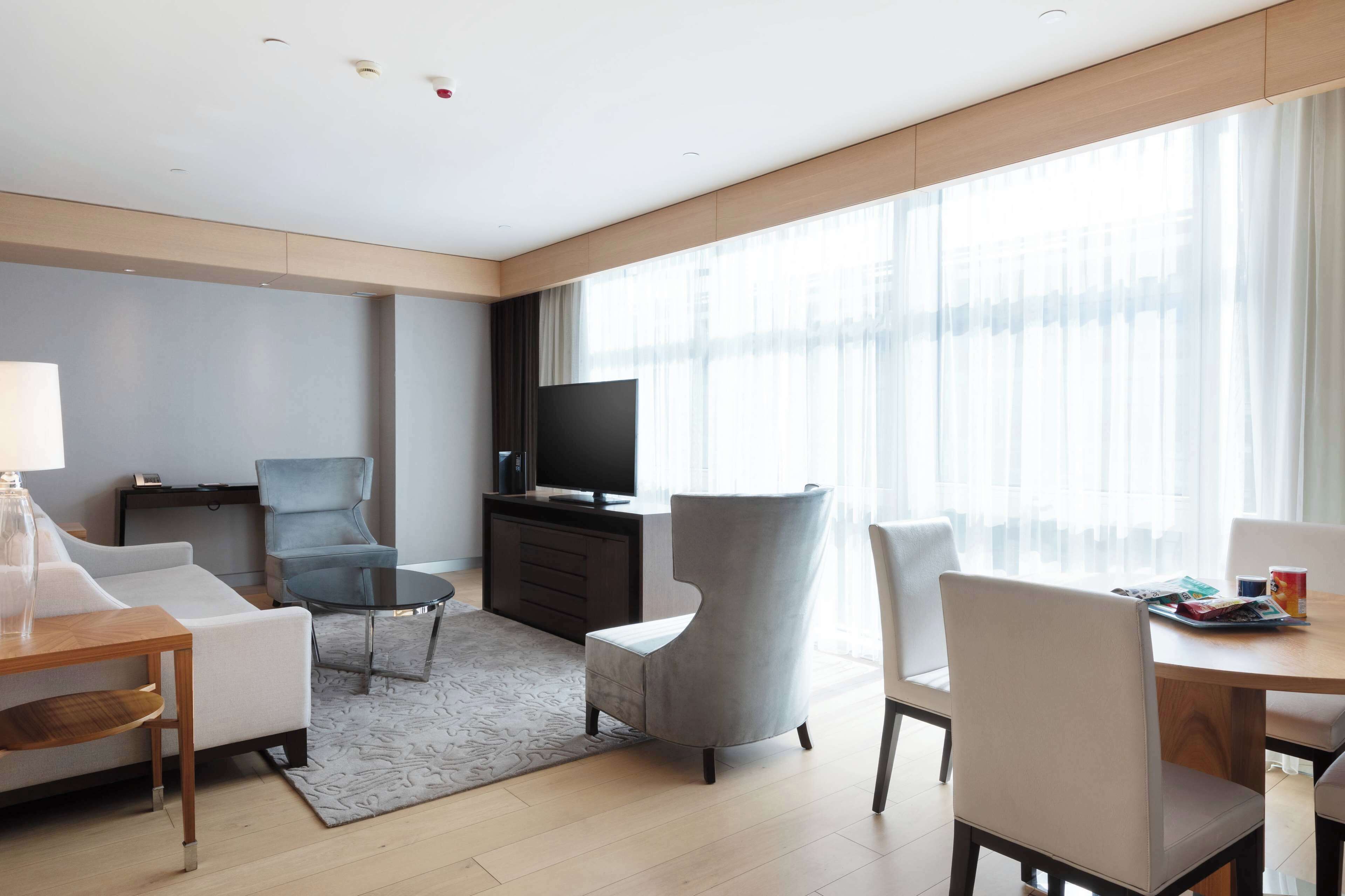 Una habitación de hotel moderna con sofá, sillones, una mesa de comedor, televisor y ventanas amplias.