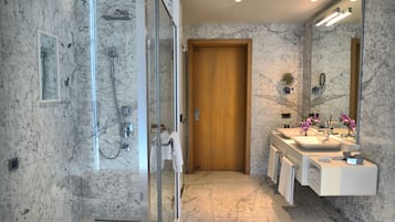 A modern bathroom with a glass-enclosed shower, marble walls, a wooden door, and a vanity with a mirror and sink.