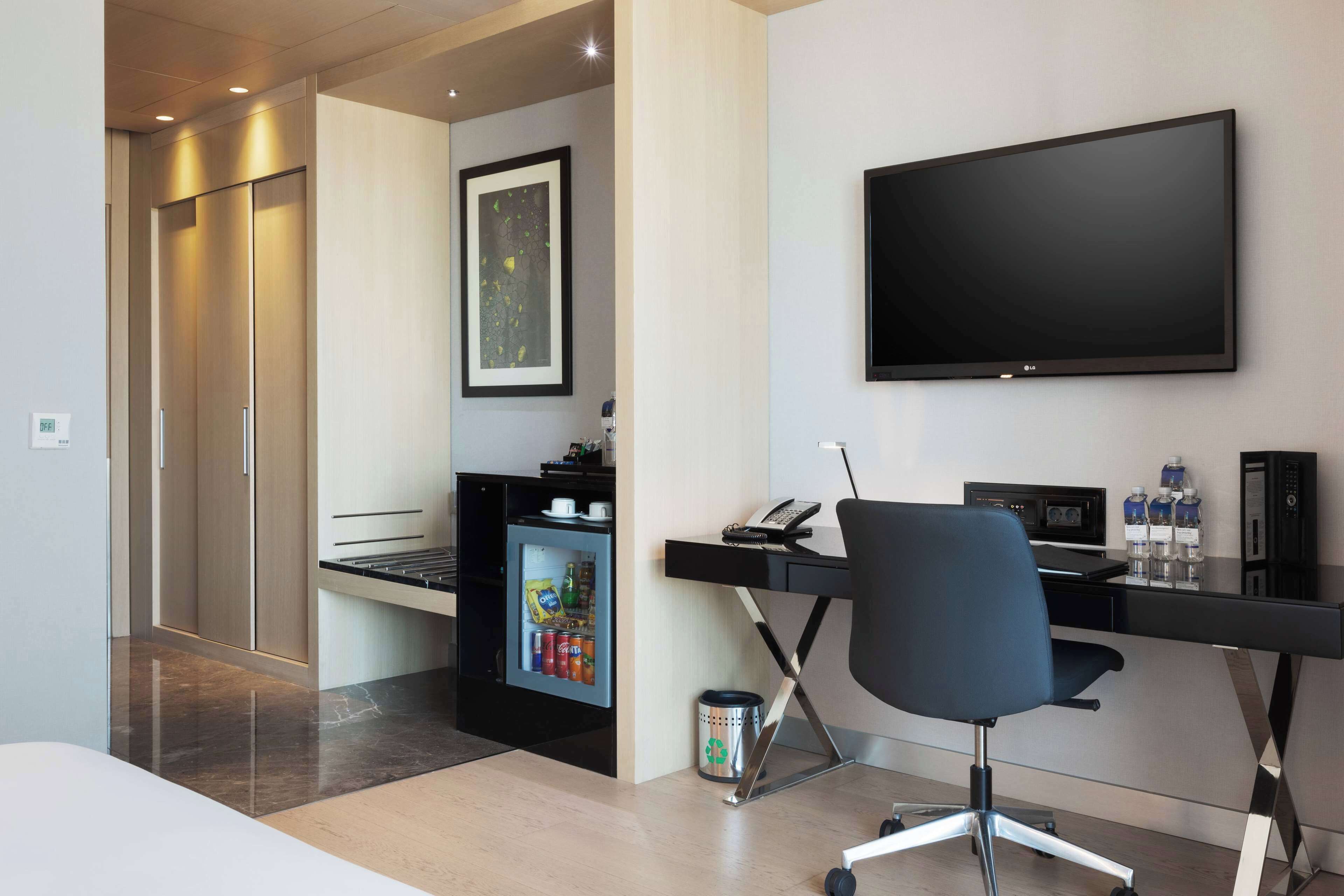 Ett modernt hotellrum med ett skrivbord, en stol, en platt-TV, en minibar och en dörr som leder ut till en korridor.