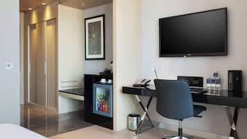 Ett modernt hotellrum med ett skrivbord, en stol, en platt-TV, en minibar och en dörr som leder ut till en korridor.