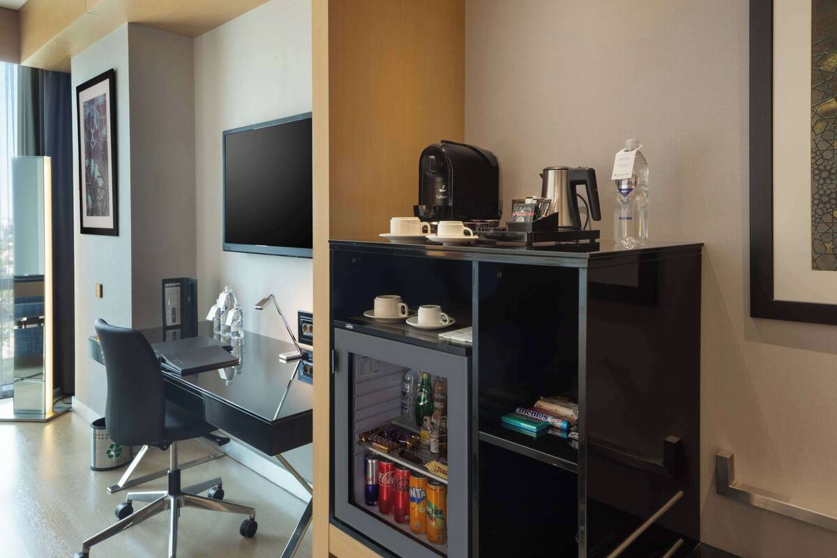 Ein modernes Hotelzimmer mit Schreibtisch, Stuhl, Kaffeemaschine und einer Minibar mit Getränken.