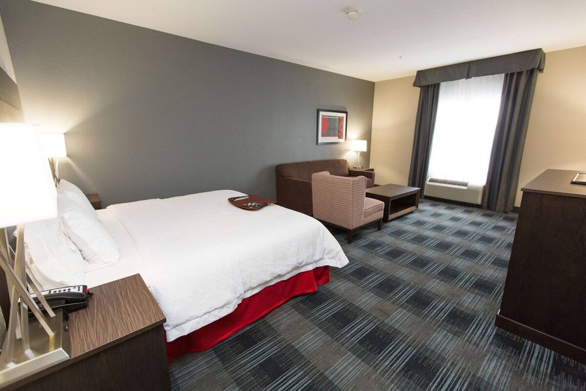 A hotel room with a bed, a desk, a chair, a lamp, a picture on the wall, and a window with curtains.