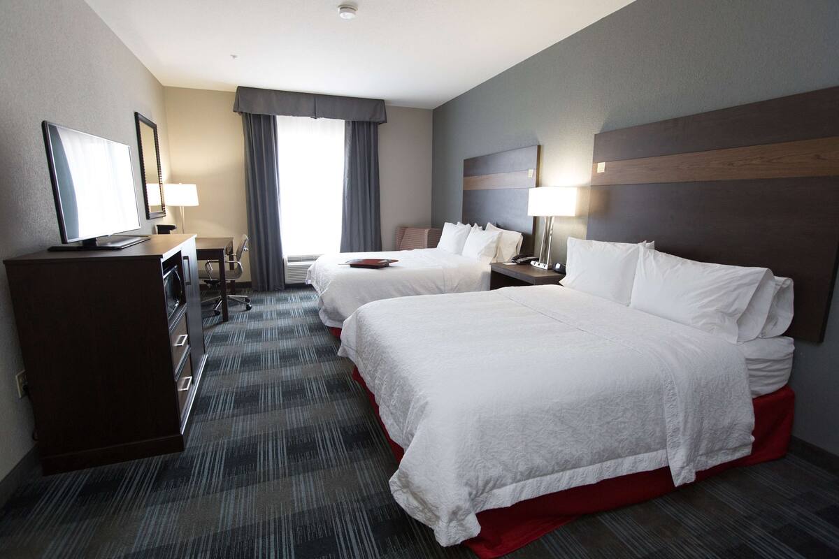 A hotel room with two beds, a desk, a TV, and a window with curtains.