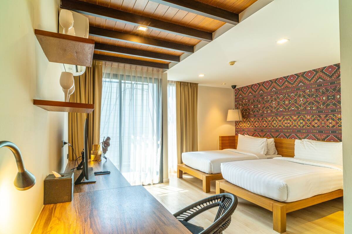 A hotel room with two beds, a desk with a lamp, a chair, and a patterned wall.