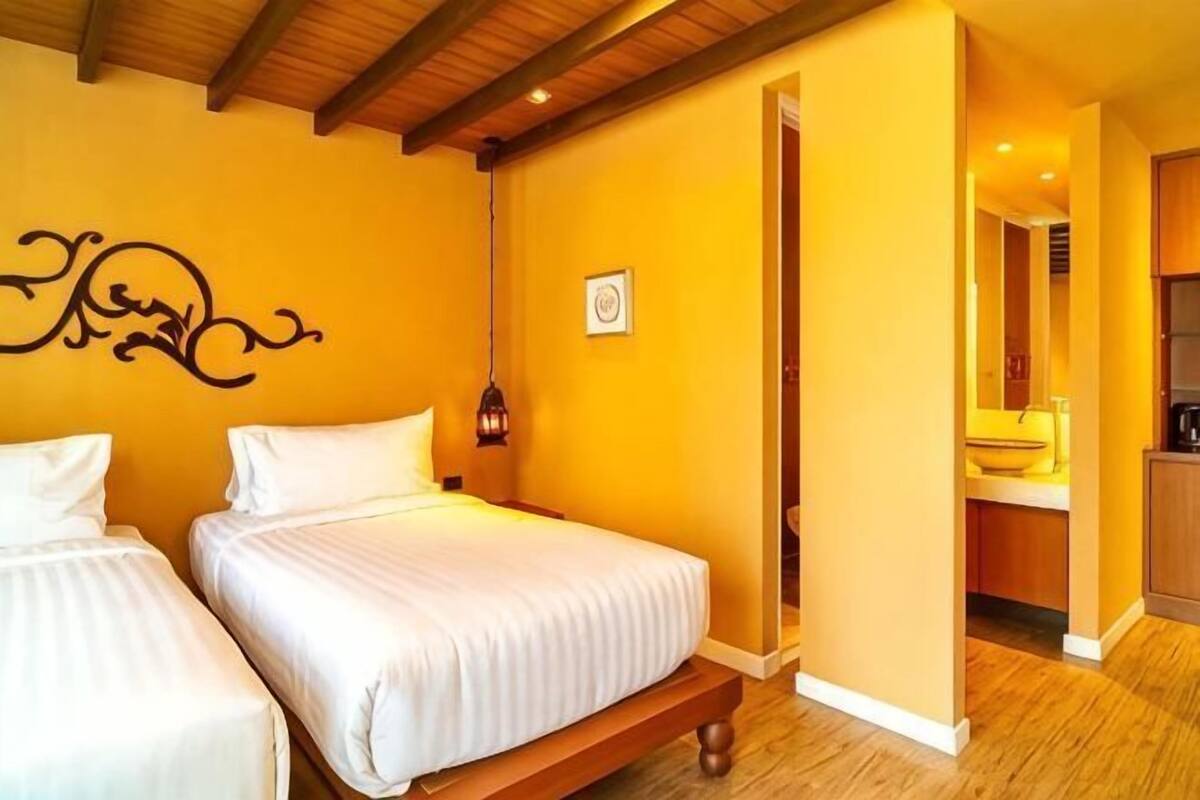 A room with two beds, a wooden ceiling, a yellow wall, and a bathroom visible through an open door.