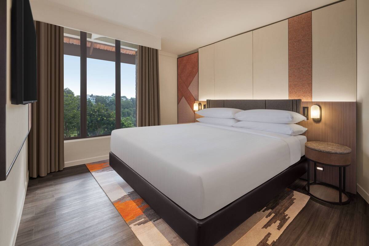 A modern hotel room with a large bed, a nightstand, a TV mounted on the wall, and a view of the outdoors through the window.