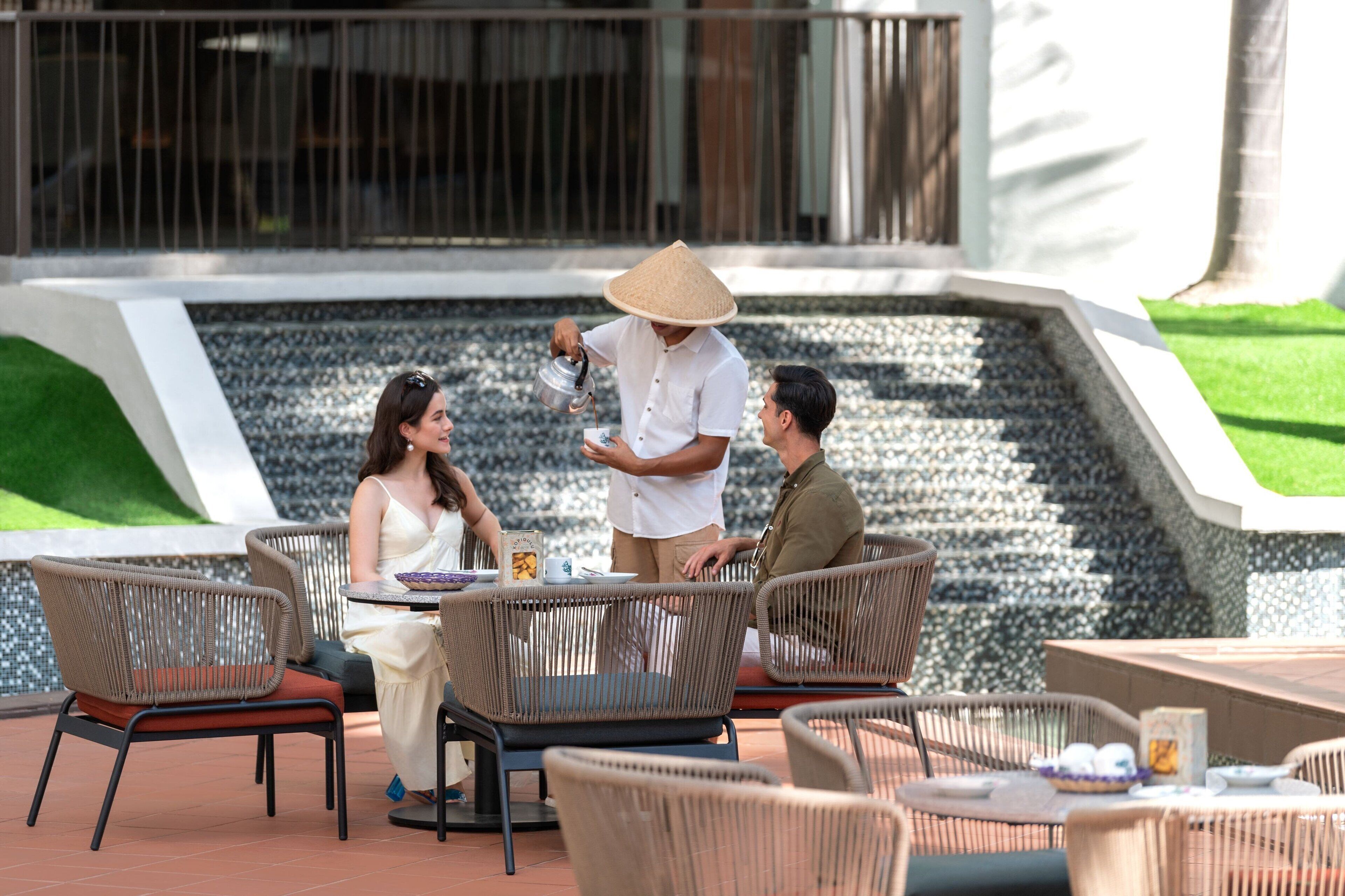 An outdoor seating area with wicker chairs and tables, a water feature in the background, and a modern building facade.