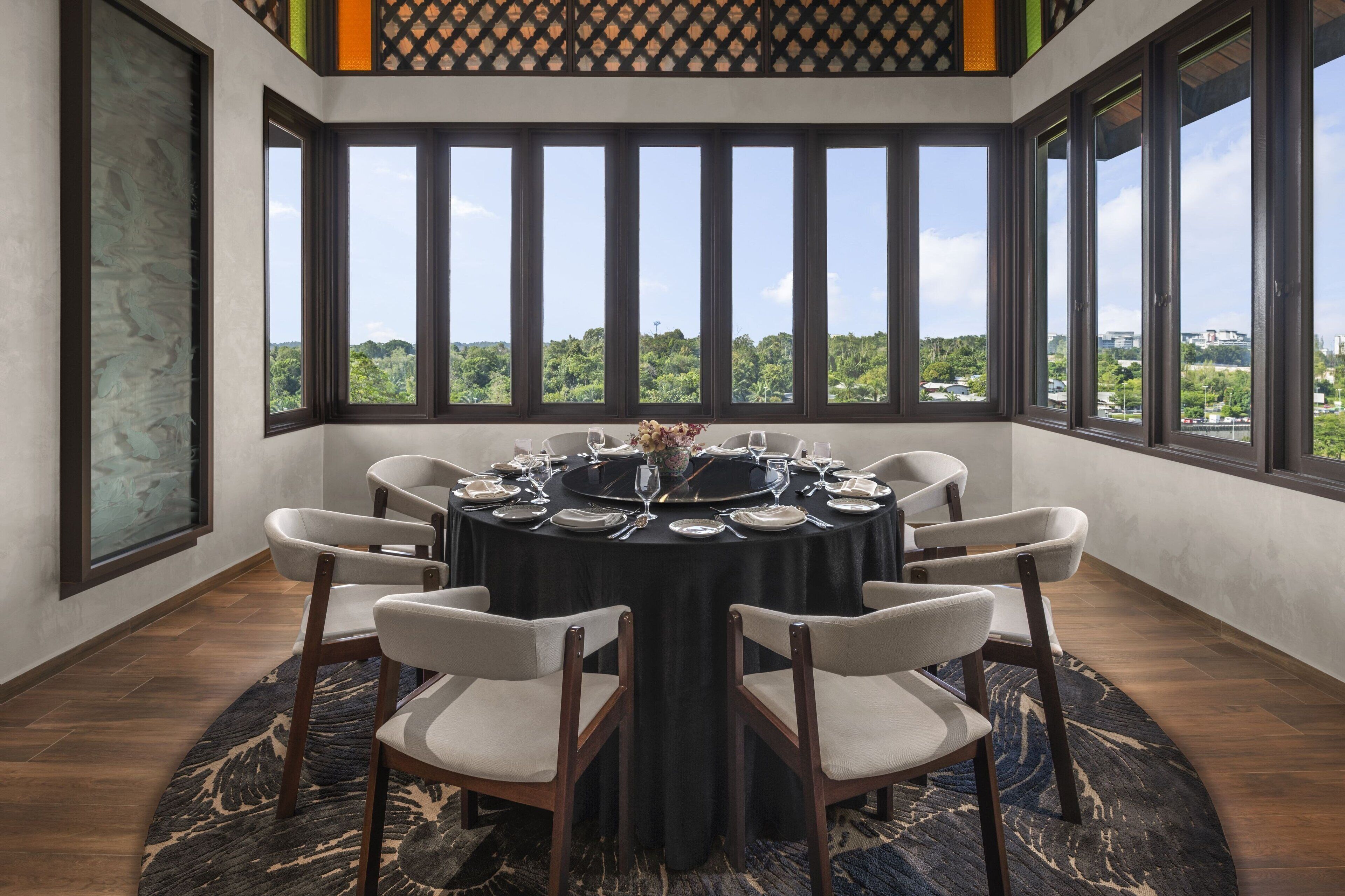 A dining area with a round table set for six, surrounded by six chairs, large windows offering a view of greenery and buildings, and a patterned rug on the floor.