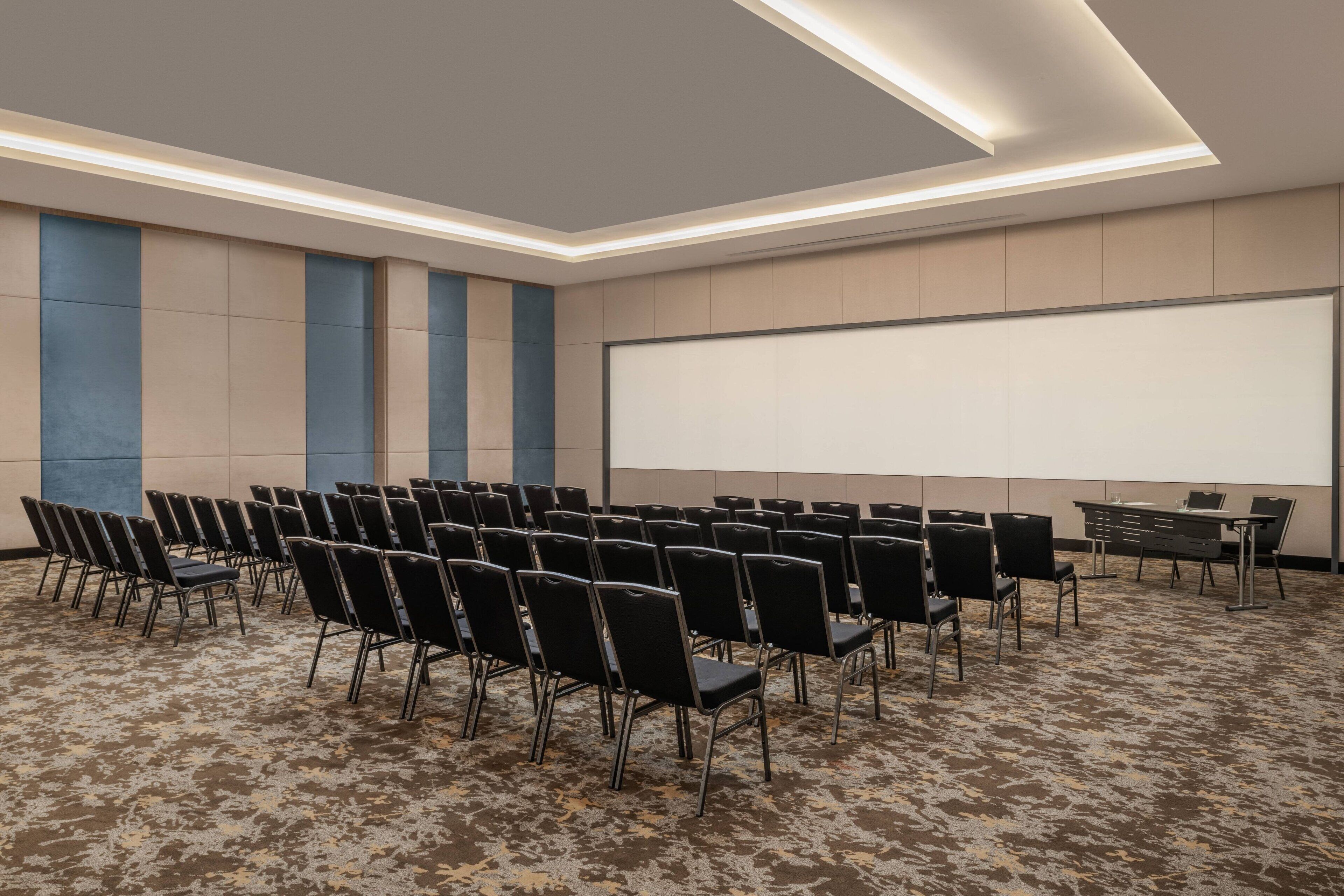 A conference room with rows of black chairs, a large projection screen, and a carpeted floor.