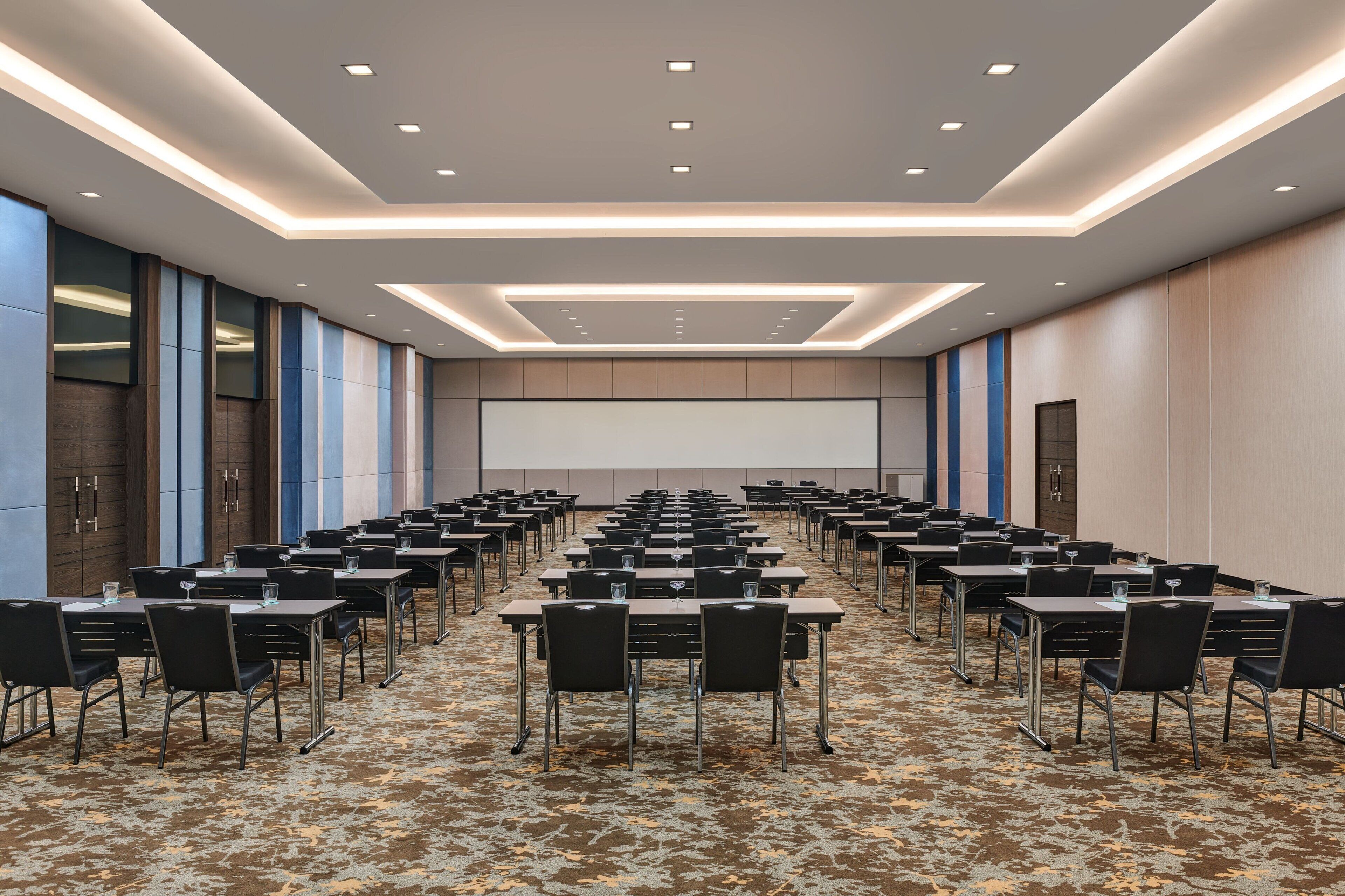A conference room with rows of tables and chairs, a large screen at the front, and a patterned carpet.