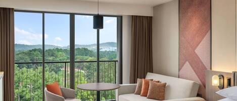 A hotel room with a large window offering a scenic view of greenery and hills. There is a sofa, a chair, and a bed with a bedside table.