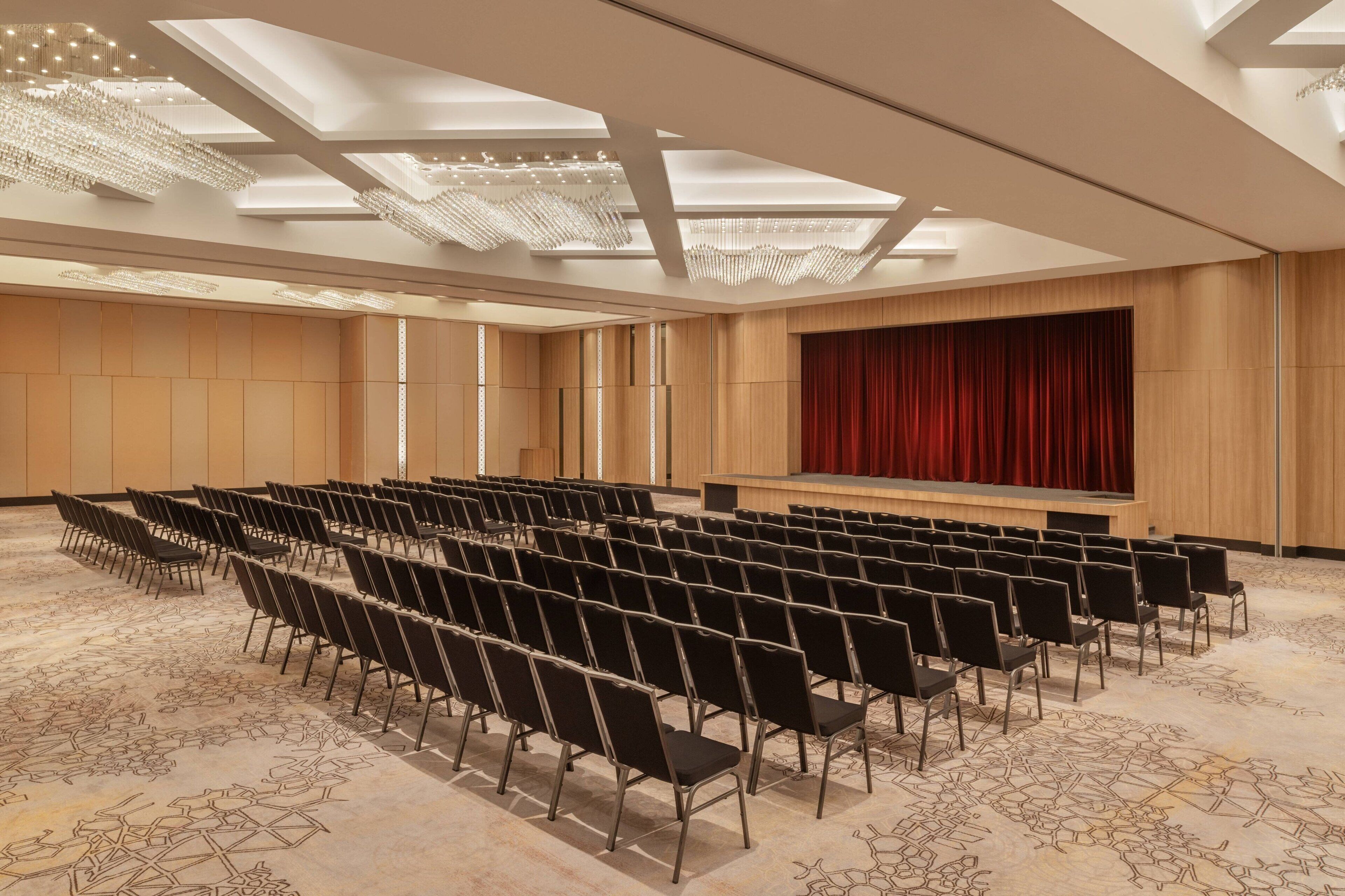 Une salle de conférence avec des rangées de chaises, une scène surmontée d’un rideau rouge et un plafond agrémenté de lucarnes et de lustres.