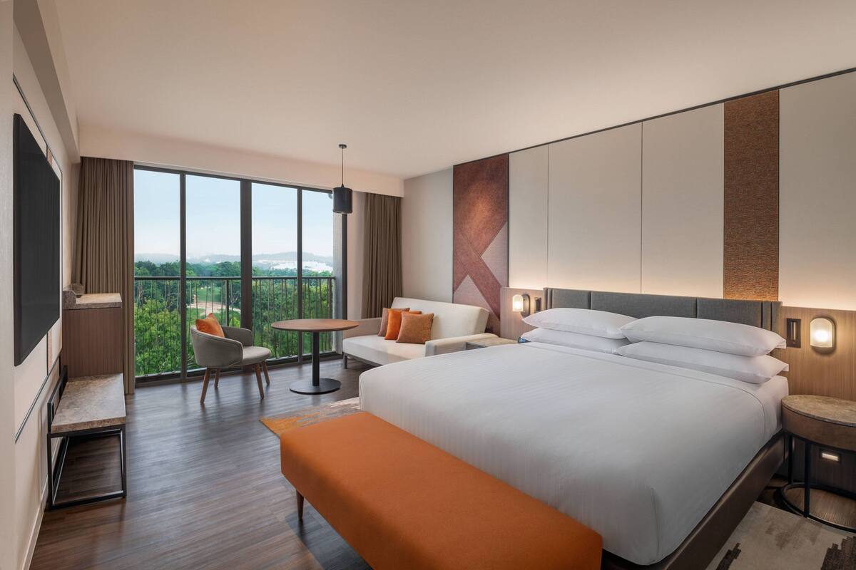 A modern hotel room with a large bed, a seating area with a table, and a view of greenery.