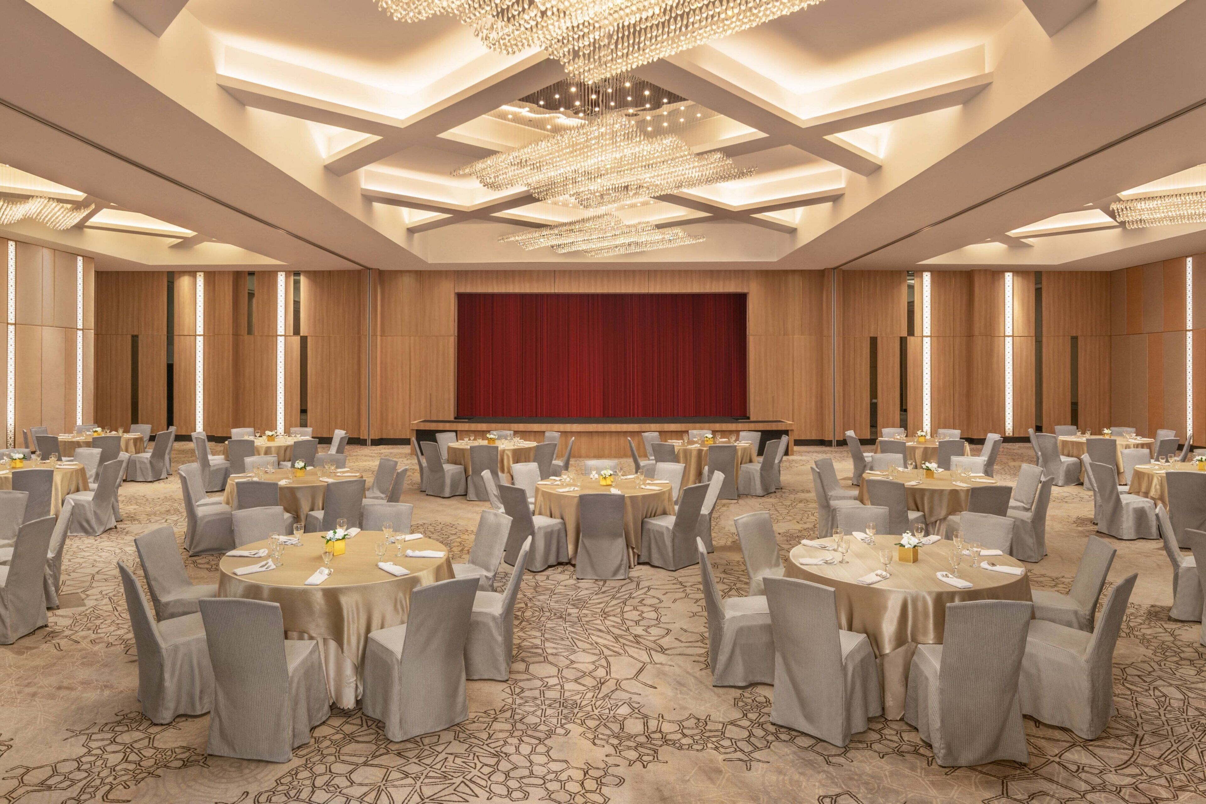 Une salle de banquet avec des tables rondes, des chaises grises et une grande scène rouge.