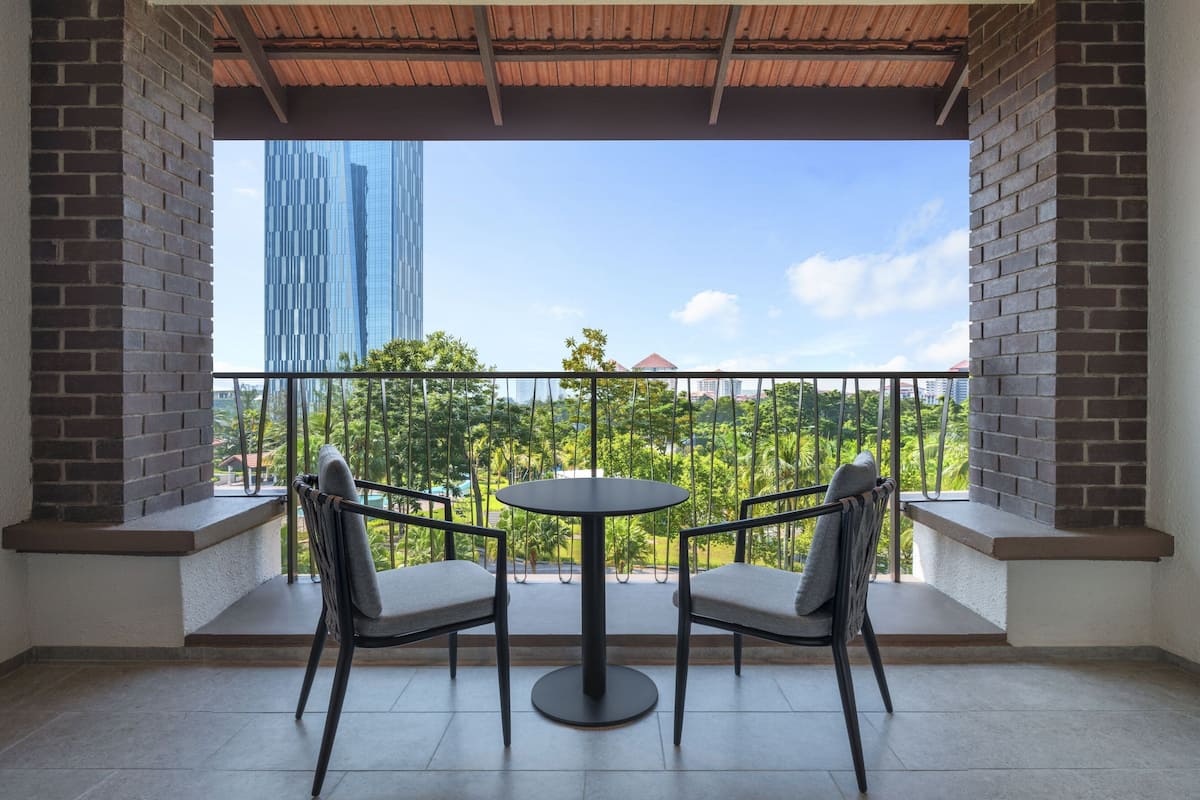 A balcony with a round table and two chairs, overlooking a cityscape with a tall building and greenery.