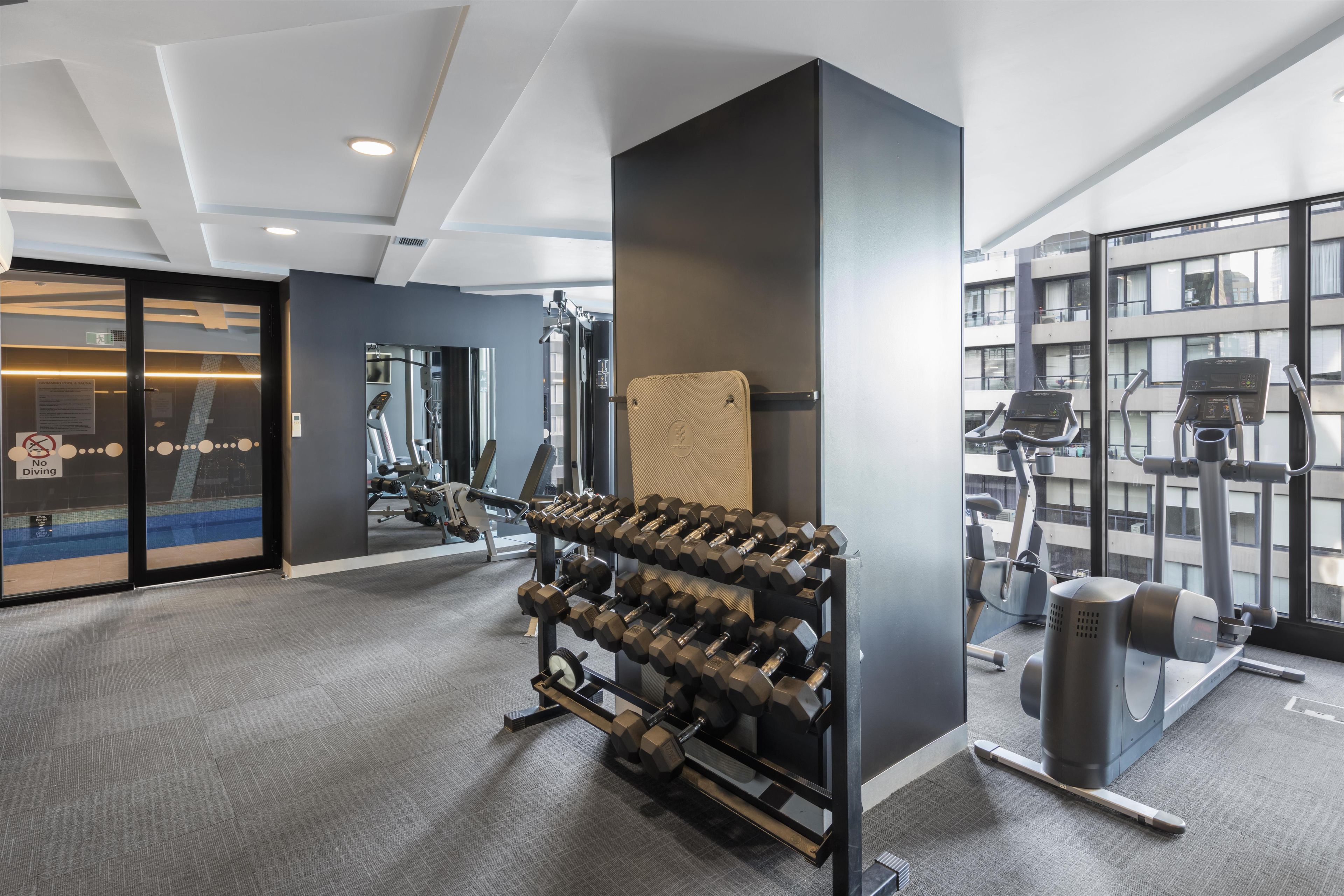 A gym with exercise equipment, a dumbbell rack, and a large window with a view of buildings.